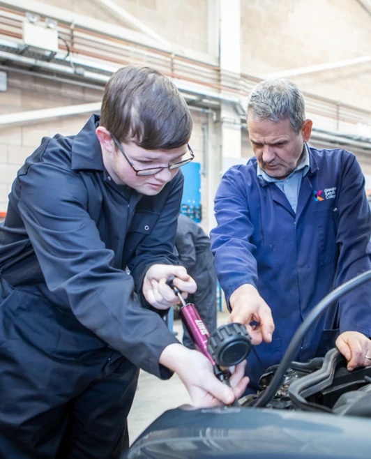 Instructor assisting a student as they use diagnostic equipment under the hood of a car in an automotive workshop. Instructor assisting a student as they use diagnostic equipment under the hood of a car in an automotive workshop.